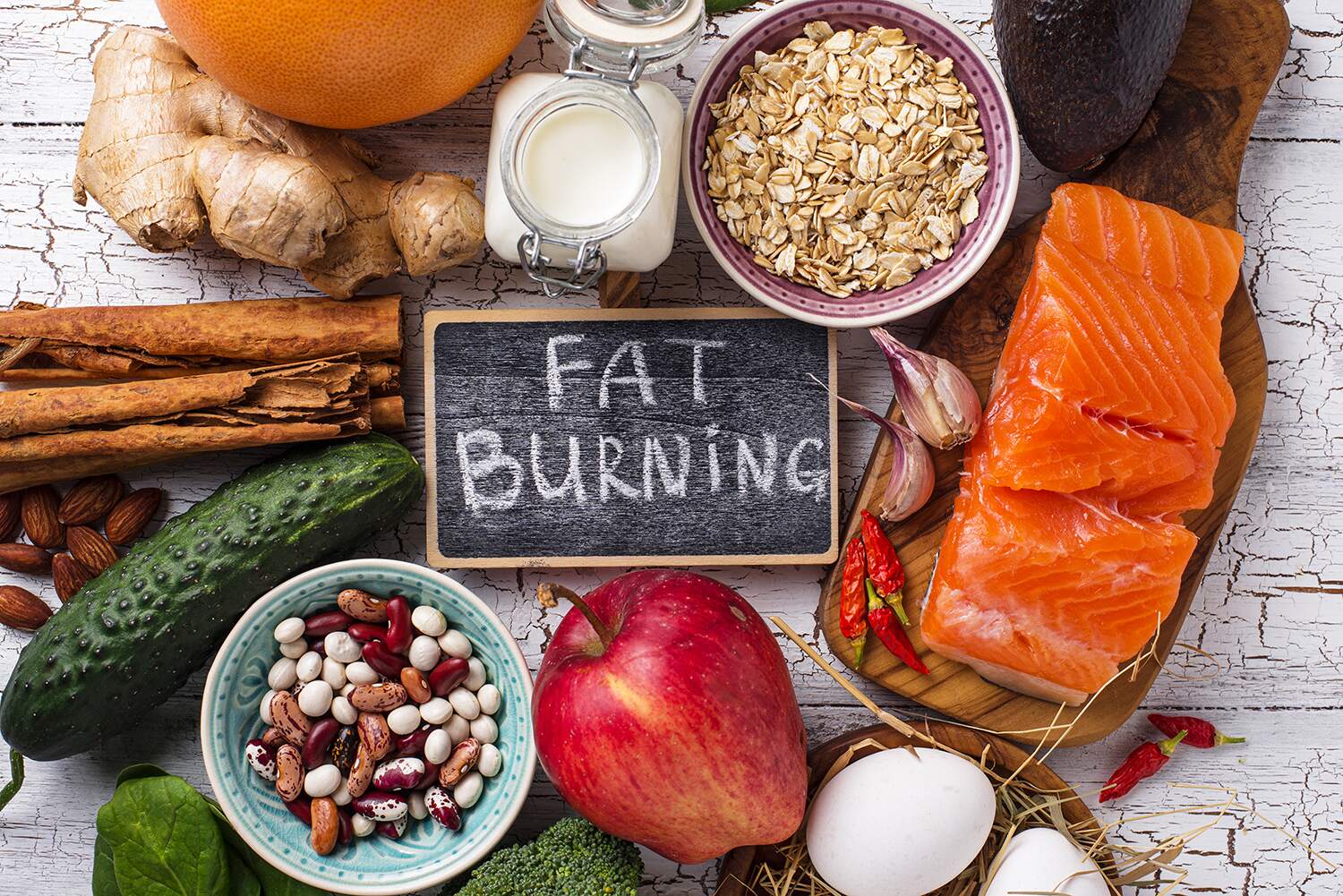 Assortment of whole foods associated with fat burning including salmon, avocado, eggs, beans, fruit and spices arranged around a chalkboard reading 'FAT BURNING' on a rustic wooden surface